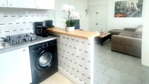 A compact kitchen with a washing machine, a coffee maker, and a sink. There is a small dining area with a table and a sofa. A painting of a cityscape is on the wall. - Spacious and bright, a real "coup de coeur" near the metro and RER station (La Courneuve)