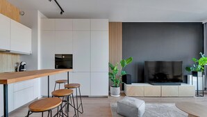 A modern kitchen with white cabinets, a wooden bar counter, and stools. There is a large flat-screen TV on a wooden stand, and a potted plant in the corner. - Brabank 20 by Grand Apartments (Gdansk)