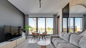 A modern living room with a large TV, a grey sofa, a dining table with chairs, and a view of construction cranes outside. - Brabank 20 by Grand Apartments (Gdansk)