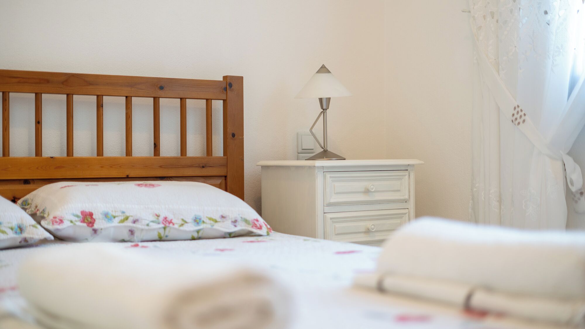 A bedroom with a wooden headboard, a bedside table with a lamp, a white dresser, and a bed with floral bedding.