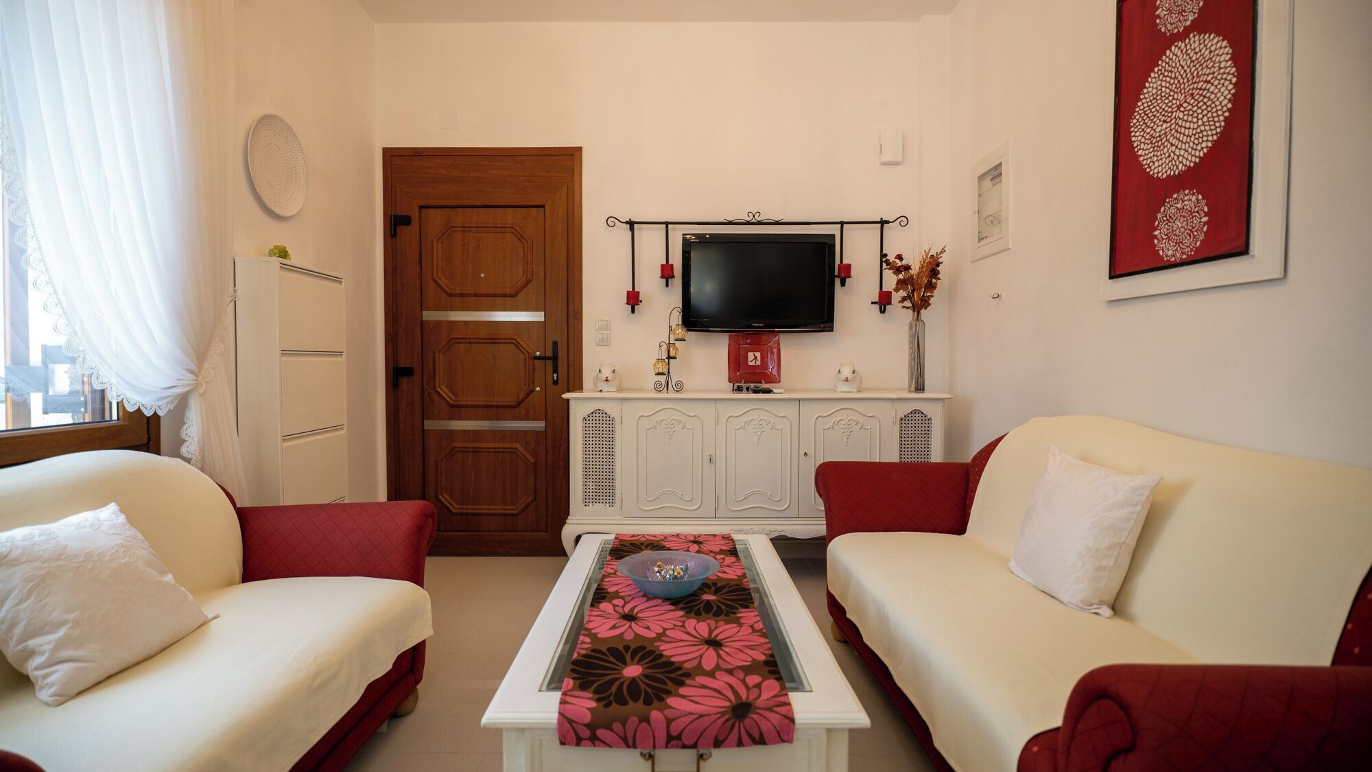 Un soggiorno con due divani color crema, un tavolino bianco con tovaglia decorata con fiori, una porta in legno, un televisore su un mobile bianco e un vaso rosso con fiori secchi.