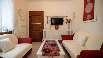 Un soggiorno con due divani color crema, un tavolino bianco con tovaglia decorata con fiori, una porta in legno, un televisore su un mobile bianco e un vaso rosso con fiori secchi.