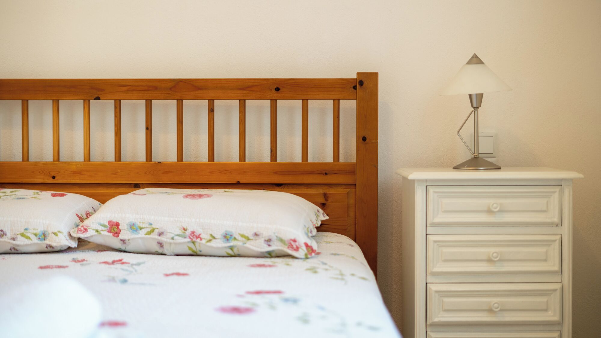 A bed with a wooden headboard, two floral pillows, and a white nightstand with a lamp and drawers.
