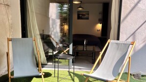 Two wooden-framed lounge chairs on artificial grass, a glass door leading to an interior space with a dining table and chairs, and a ceiling fan.