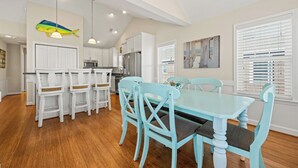 Una cocina amplia con armarios blancos, una zona de comedor con una mesa y sillas azules, y una lámpara colgante.