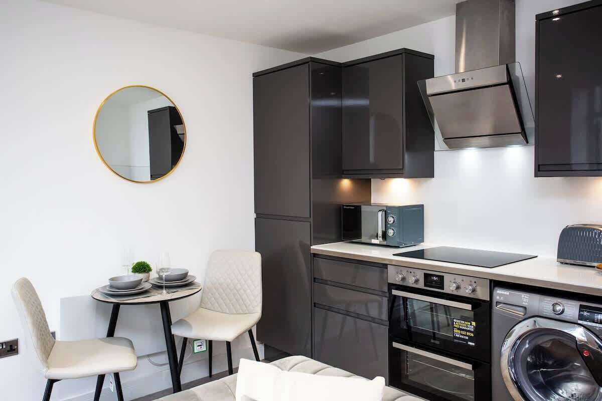Cocina compacta con microondas, horno y lavadora, un espejo redondo y una pequeña zona de comedor con sillas.