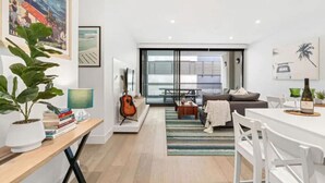 A modern living room with a sofa, coffee table, and a dining area with white chairs. There is a guitar and a plant on a side table. - Anchors Away | Escape in the heart of Mornington (Mornington)