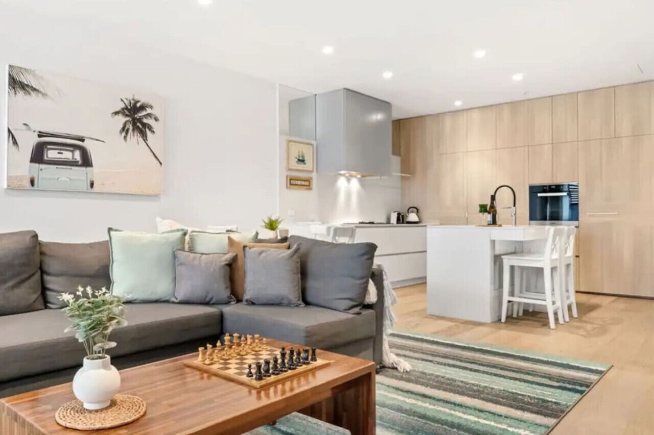 A modern living room with a sofa, a coffee table with a chess set, and a kitchen area with a refrigerator and a sink.