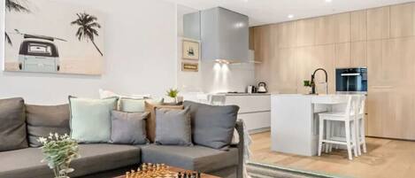 A modern living room with a sofa, a coffee table with a chess set, and a kitchen area with a refrigerator and a sink.
