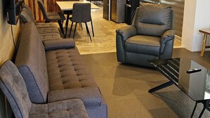 Una sala de estar moderna con sofá, sillón y una mesa de centro de vidrio. Hay un televisor montado en la pared, y se puede ver un comedor al fondo.