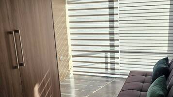 A modern living room with a dark brown wardrobe, a sofa with cushions, and a large window with blinds.