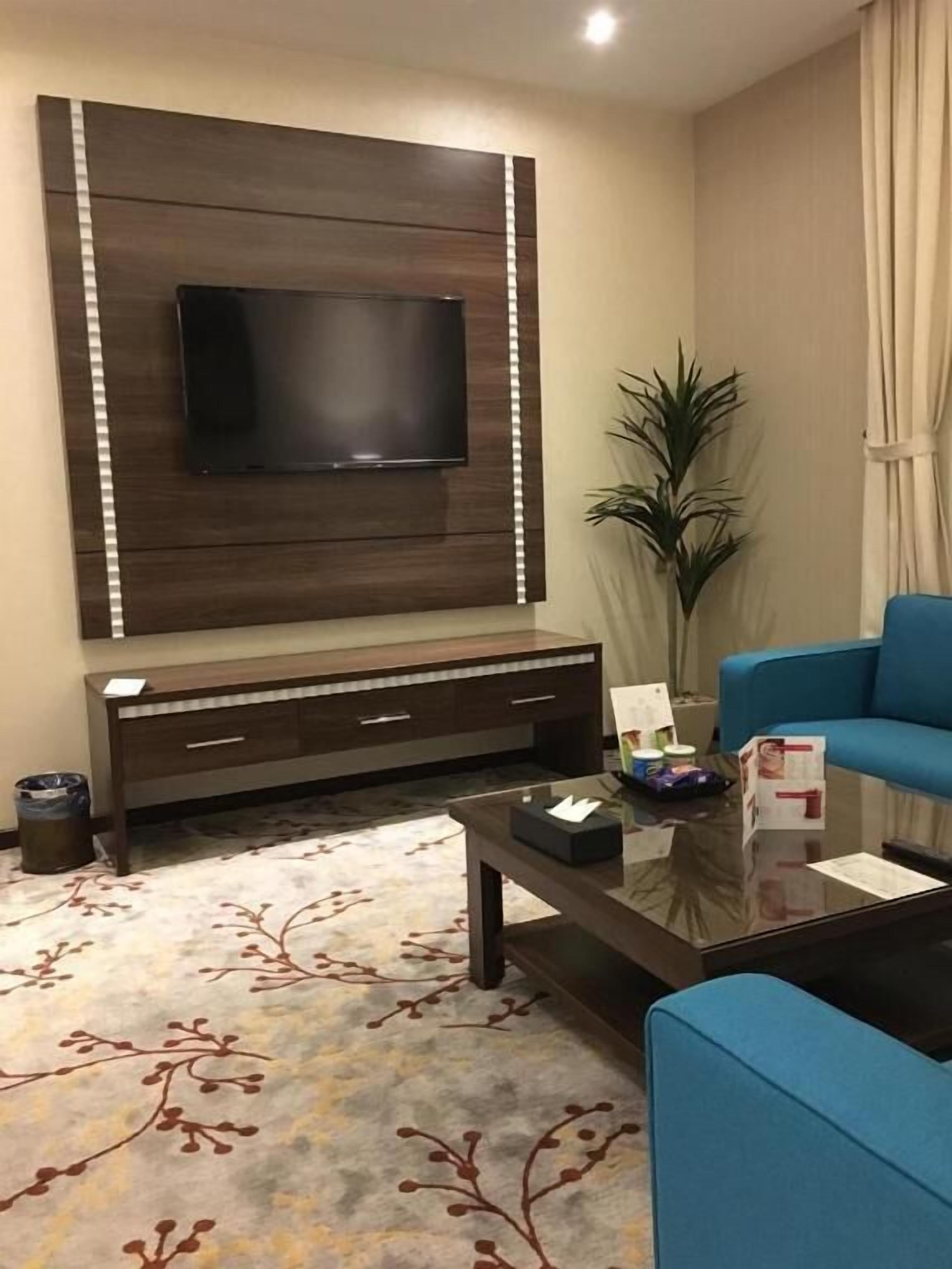 A modern living room with a flat-screen TV mounted on a wooden wall, a blue sofa, a coffee table, and a patterned rug.