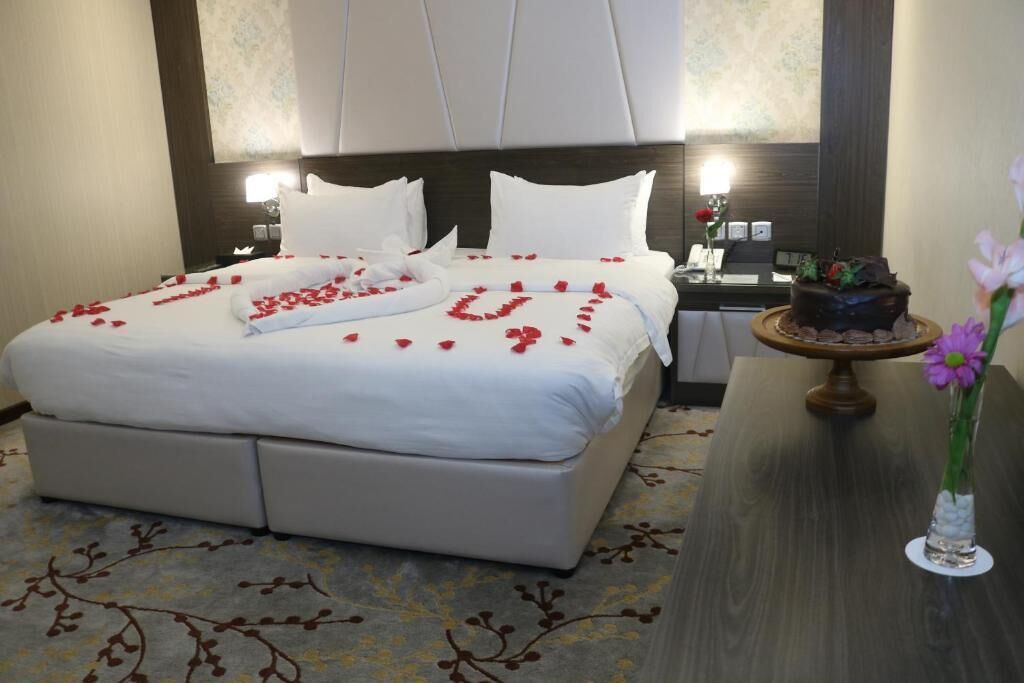 A hotel room with a bed adorned with rose petals, a chocolate cake on a wooden stand, and a vase with flowers on a table.