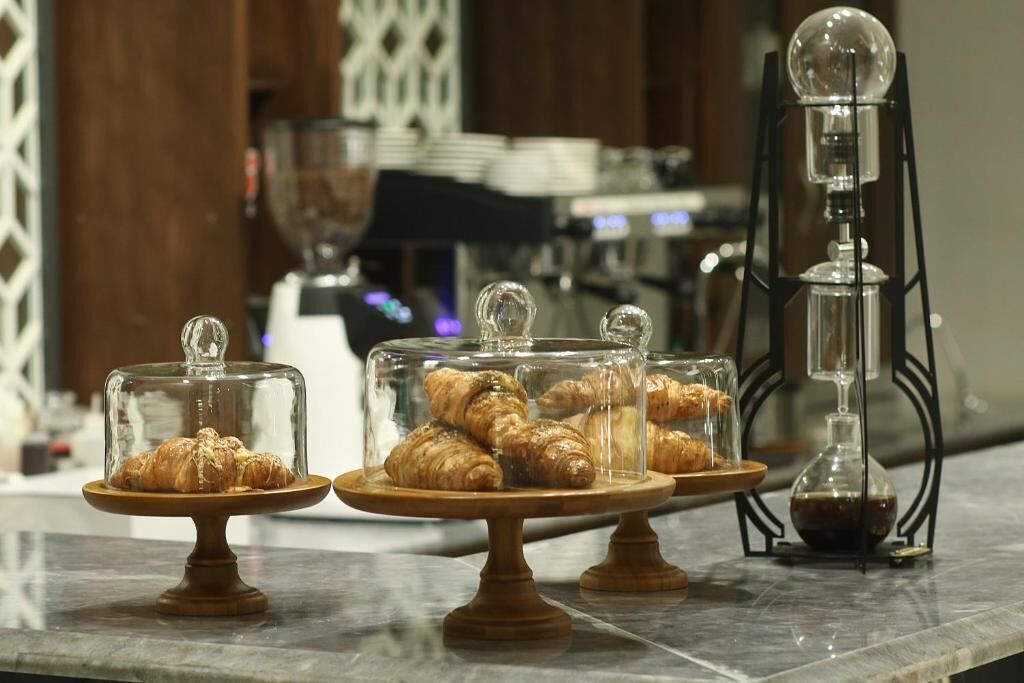 A display of pastries on wooden stands with glass domes, a coffee machine, and a decorative glass object in the background.