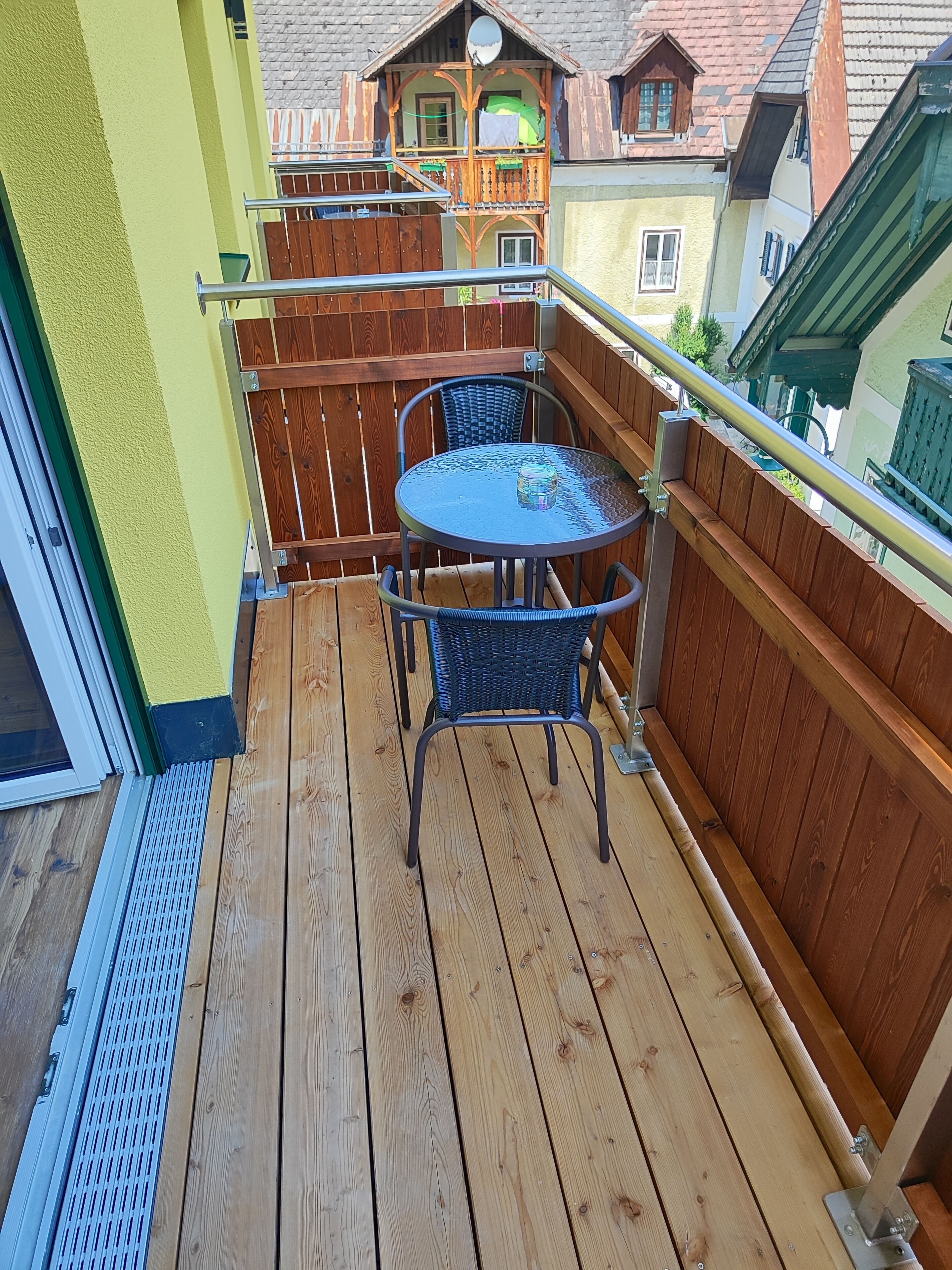 A wooden deck with a round table and two chairs, a metal railing, and a view of neighboring houses.