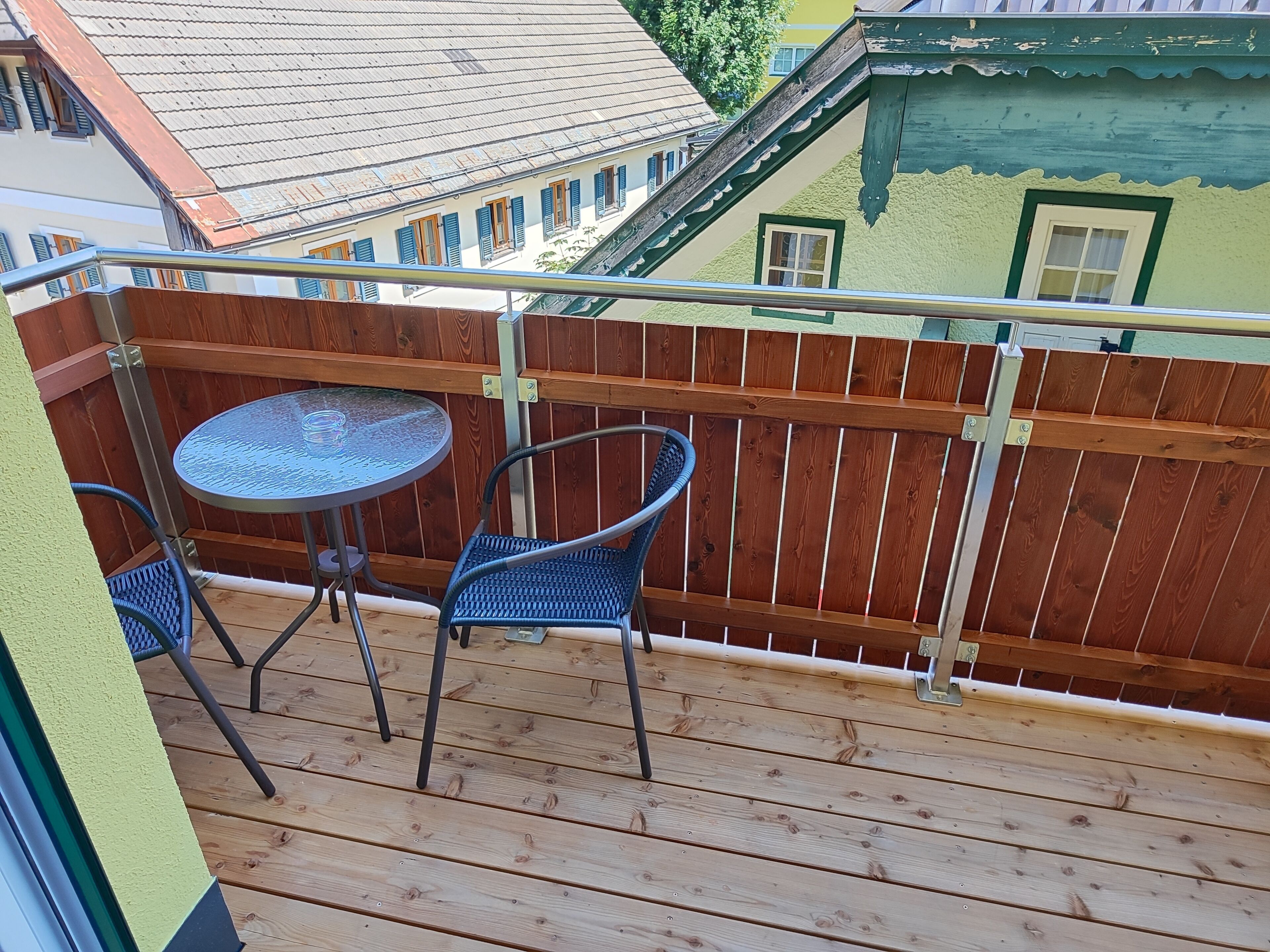 A balcony with a round table and two chairs, wooden deck, metal railing, and a view of neighboring buildings.