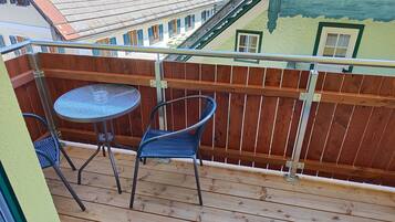 A balcony with a round table and two chairs, wooden deck, metal railing, and a view of neighboring buildings.