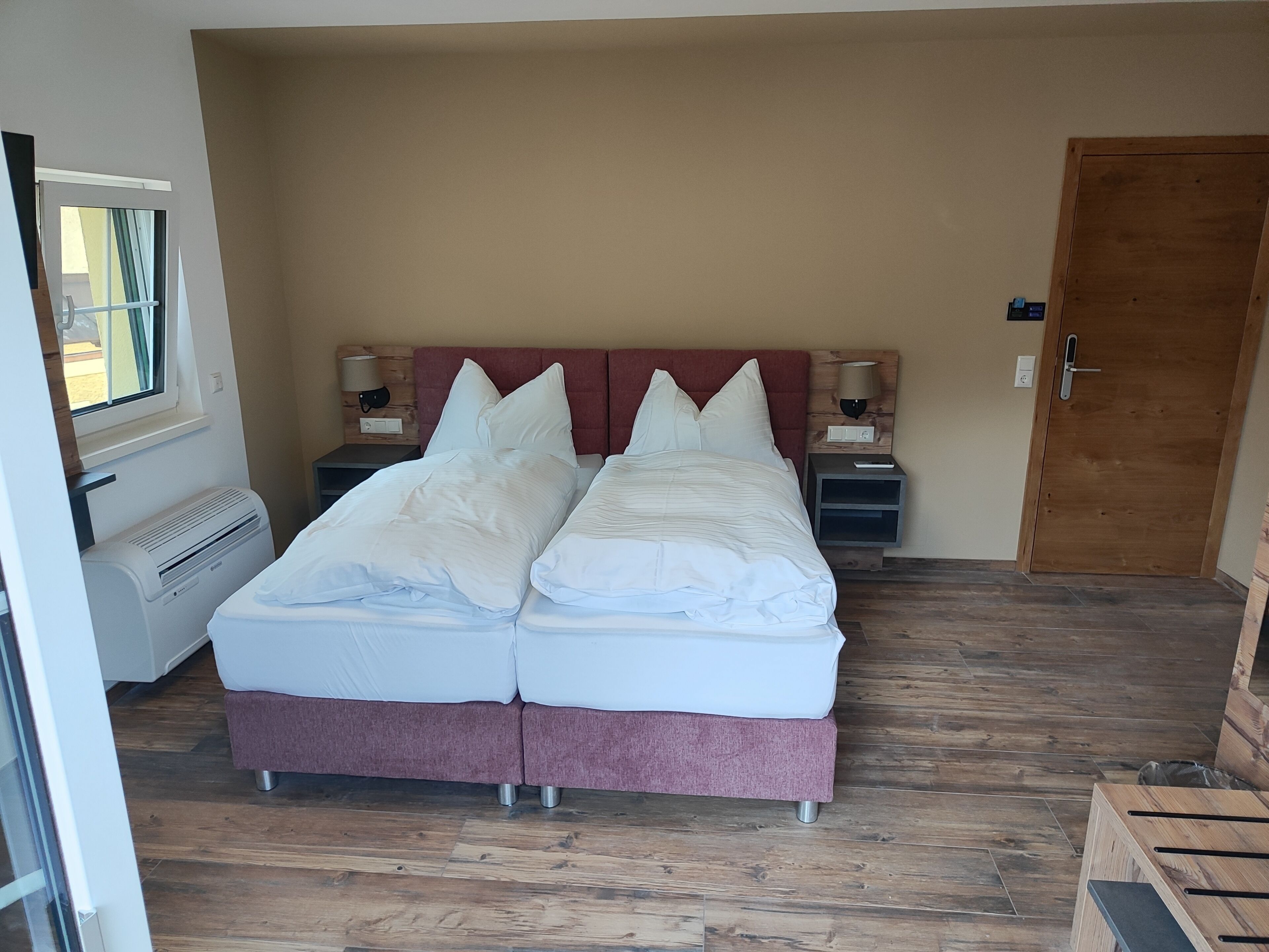 Ein Doppelbett mit weißer Bettwäsche, ein Holz-Kopfstück, zwei Nachttische mit Lampen, ein Fenster mit Vorhängen und eine Tür.