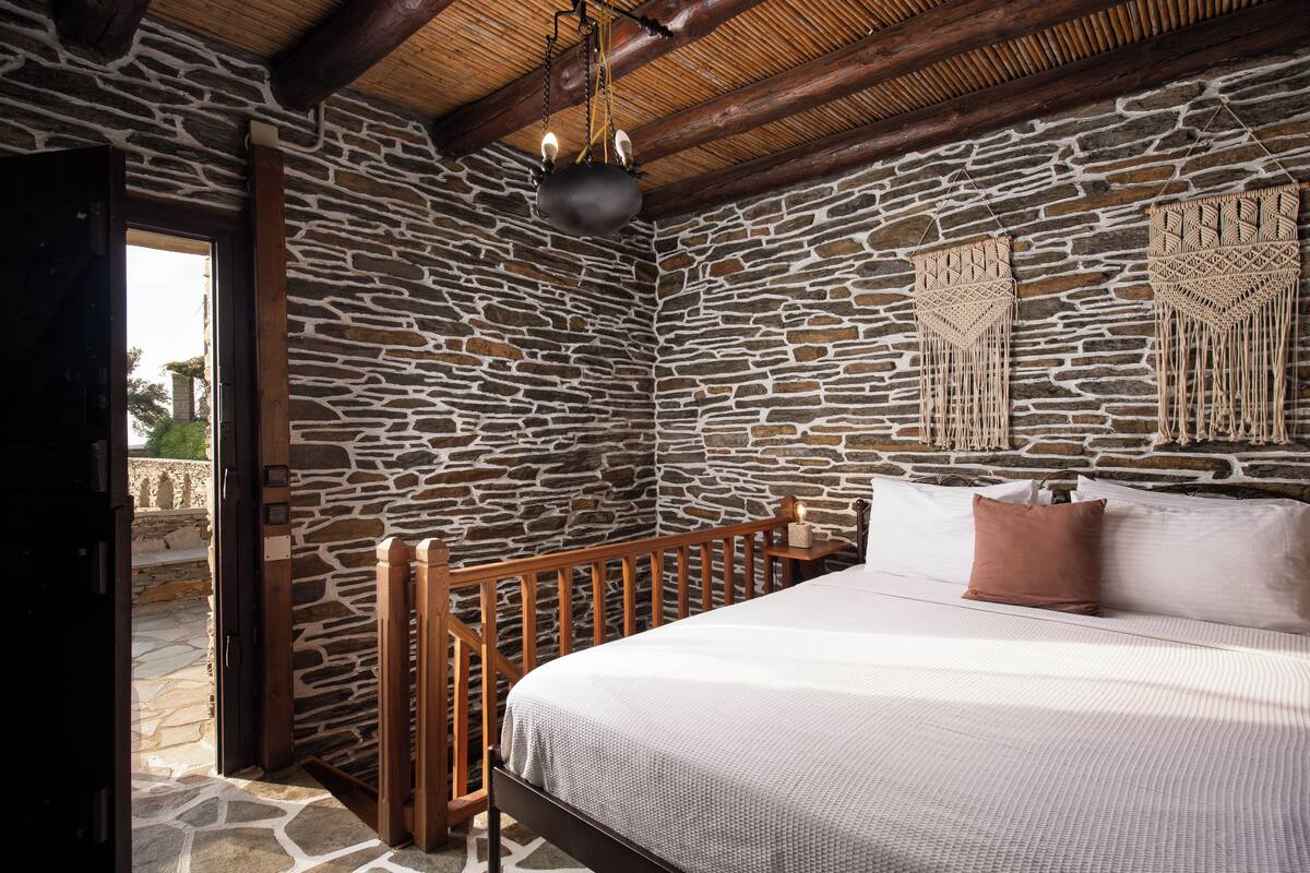 Une chambre aux murs de pierre, avec un lit, une balustrade en bois et des décorations murales en macramé.