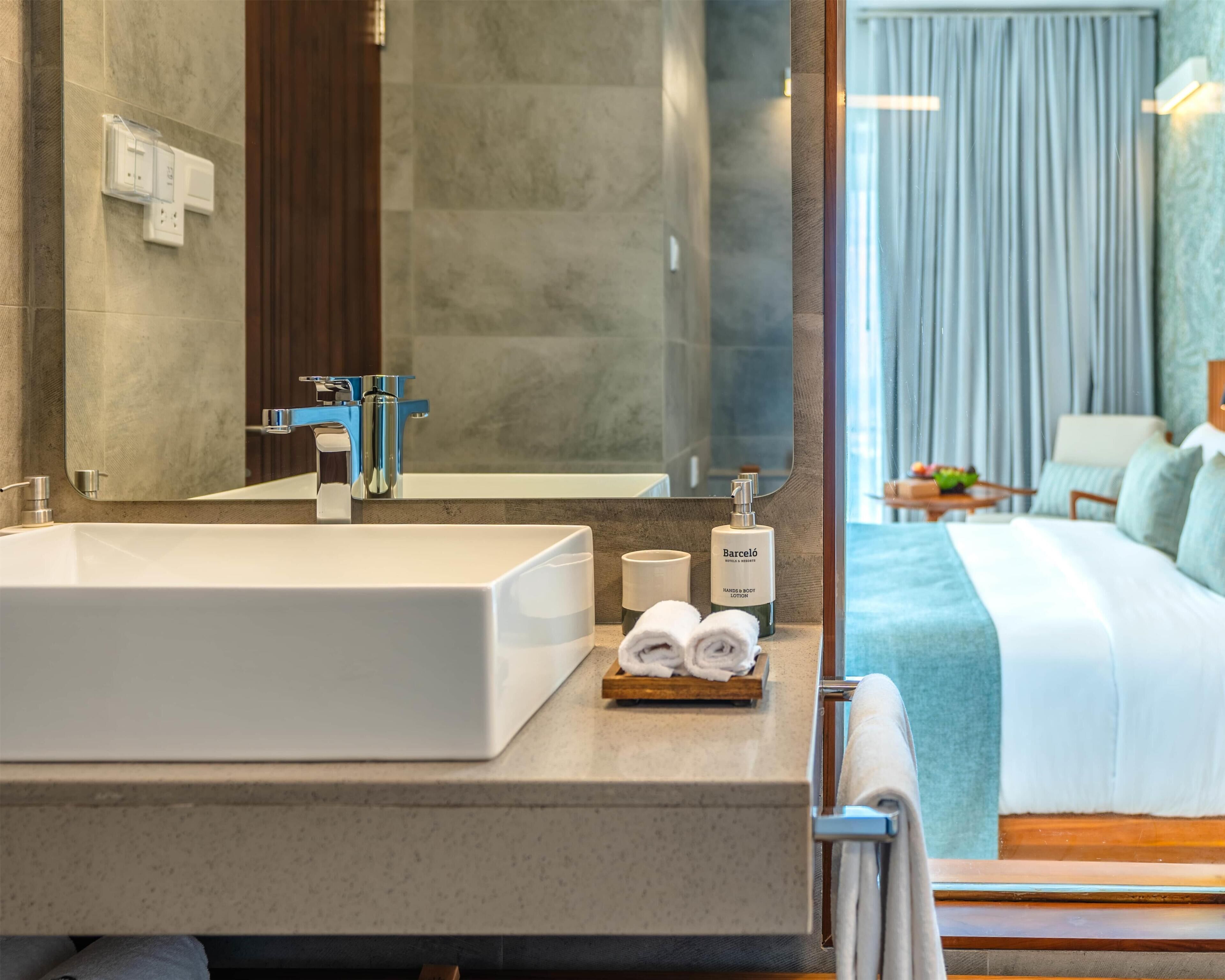 A hotel bathroom with a large white sink, a mirror, a blue faucet, a bottle of Barcelona lotion, and a towel rack with towels.