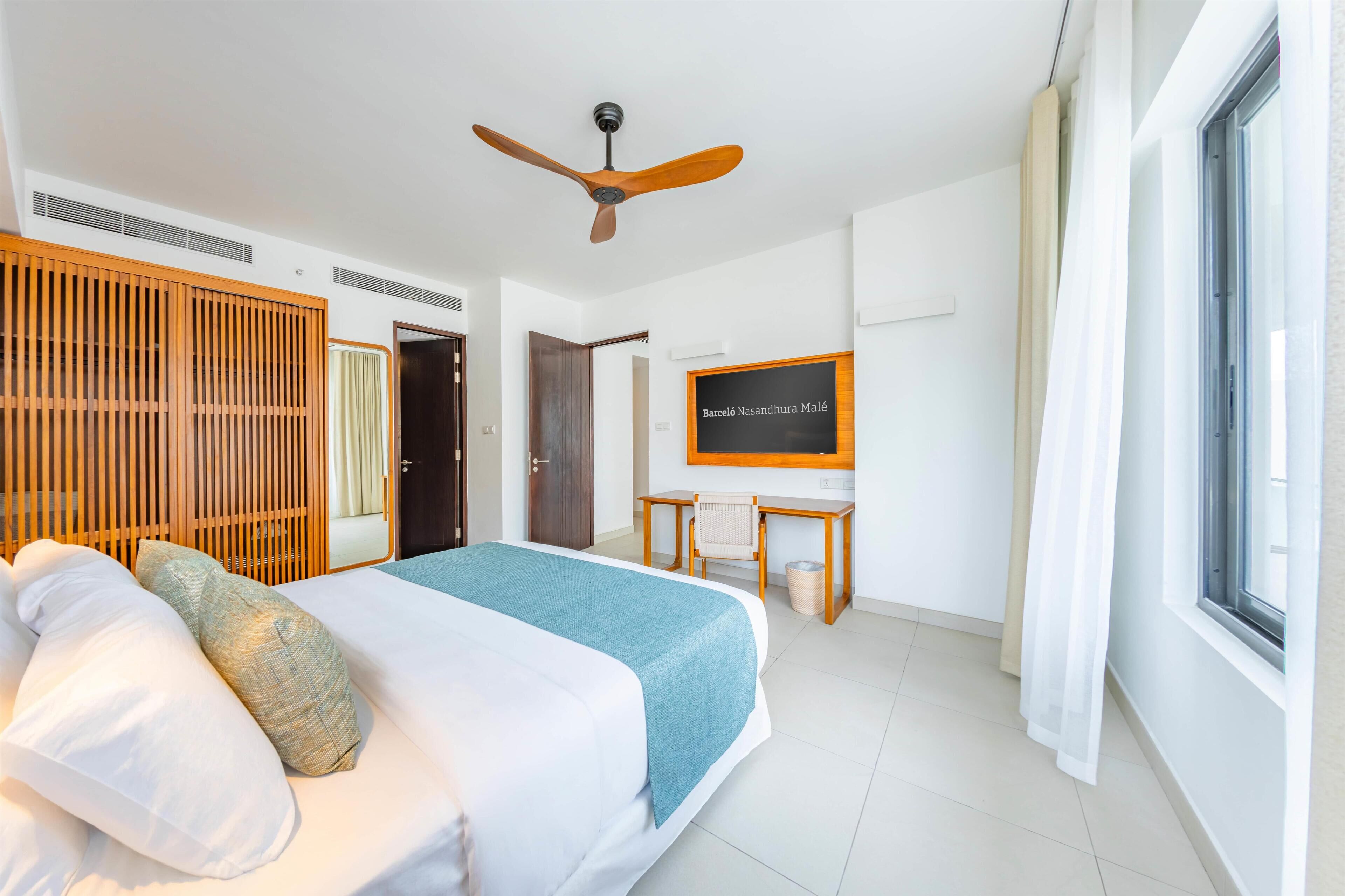 A hotel room with a bed, a desk, a ceiling fan, a window, and a wall-mounted TV.