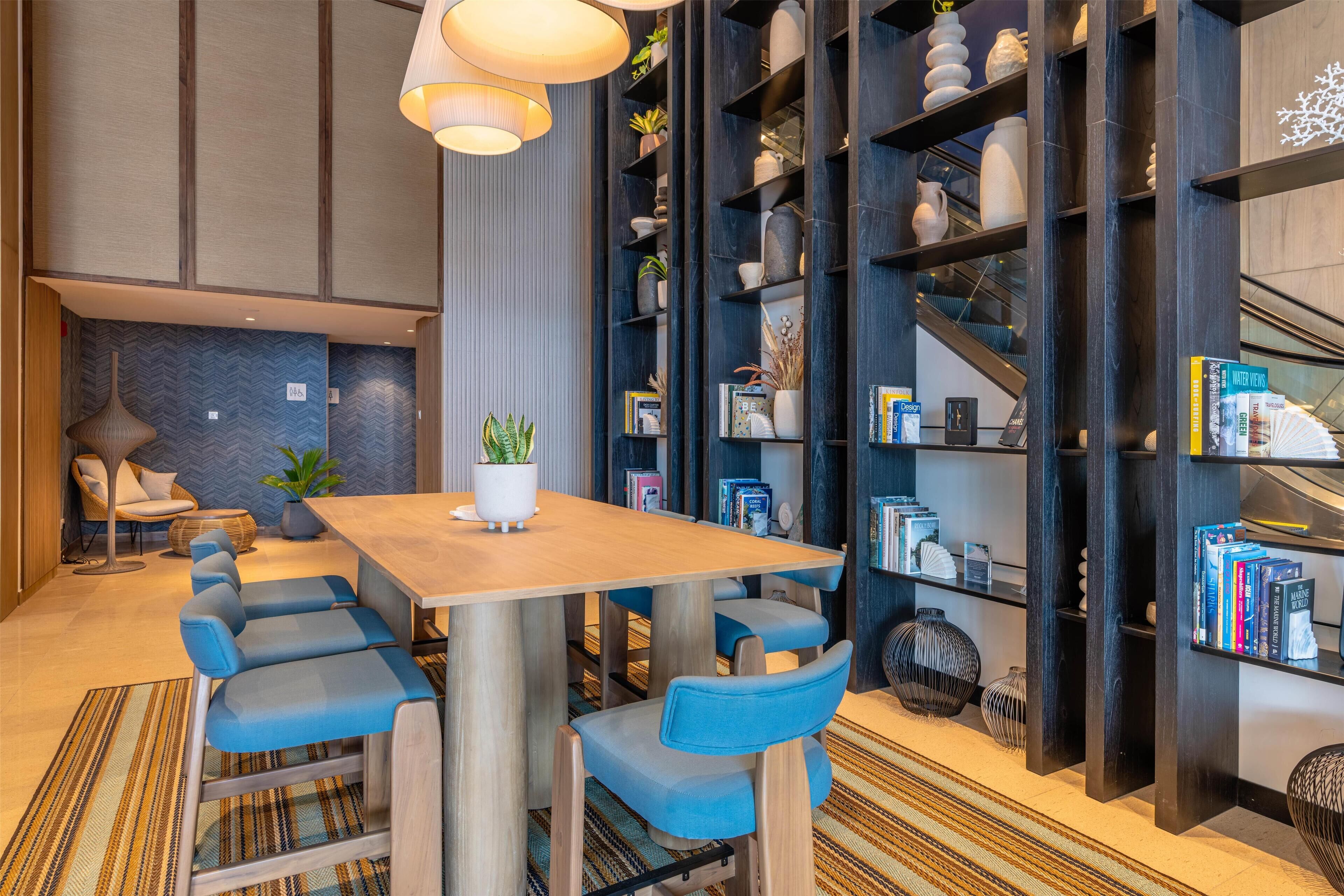 A modern interior with a wooden dining table, blue upholstered chairs, a bookshelf, and a staircase.