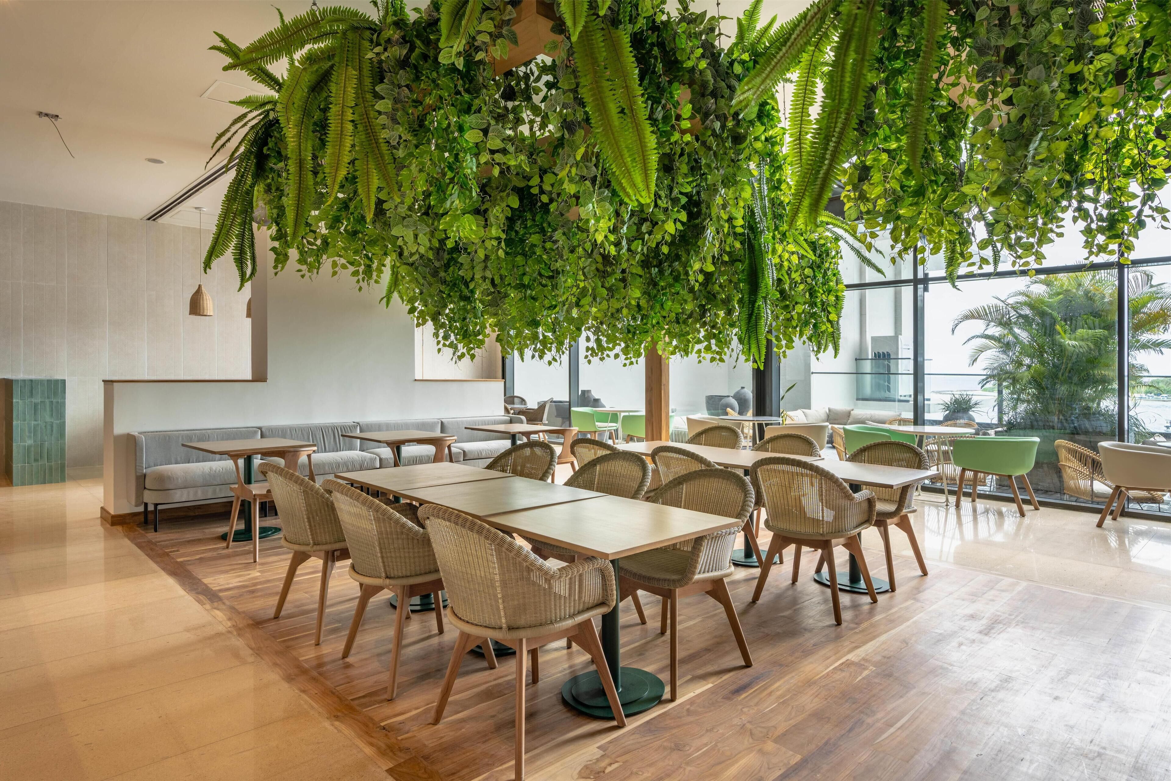 Een moderne eetruimte met houten tafels en rieten stoelen, omgeven door grote ramen en binnenplanten.