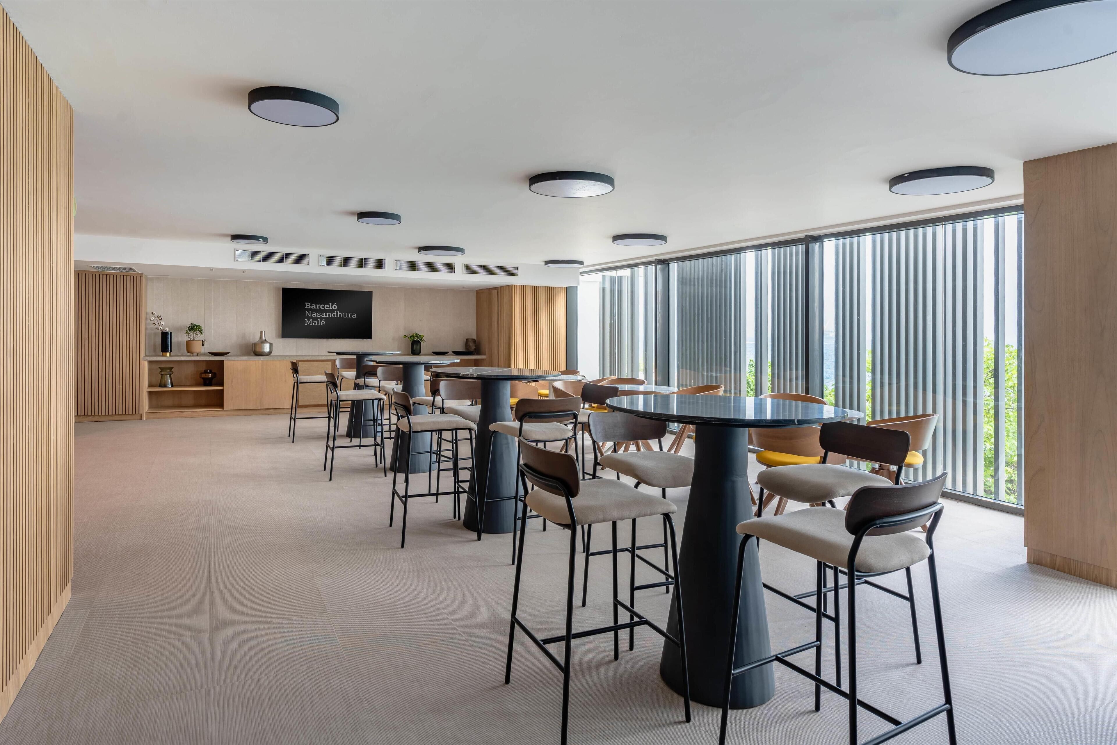 Une salle de conférence moderne, dotée de tables et de chaises rondes, d’un grand écran de télévision et de grandes fenêtres offrant une vue sur l’extérieur.