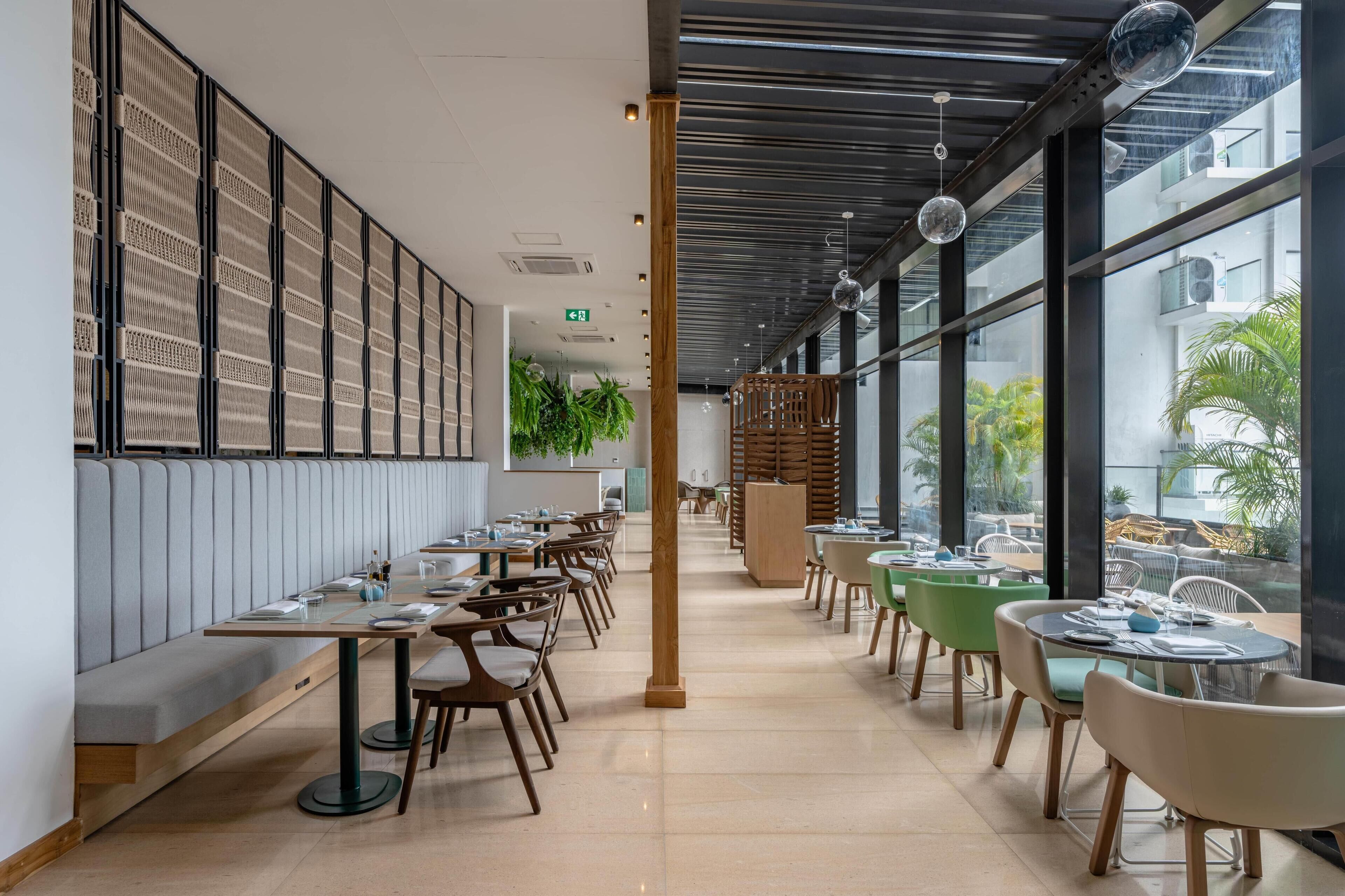 Ein modernes Restaurant mit langen Gemeinschaftstischen und Sitzecken, großen Fenstern mit Blick ins Freie und einer Decke mit freiliegenden Balken und hängenden Leuchten.
