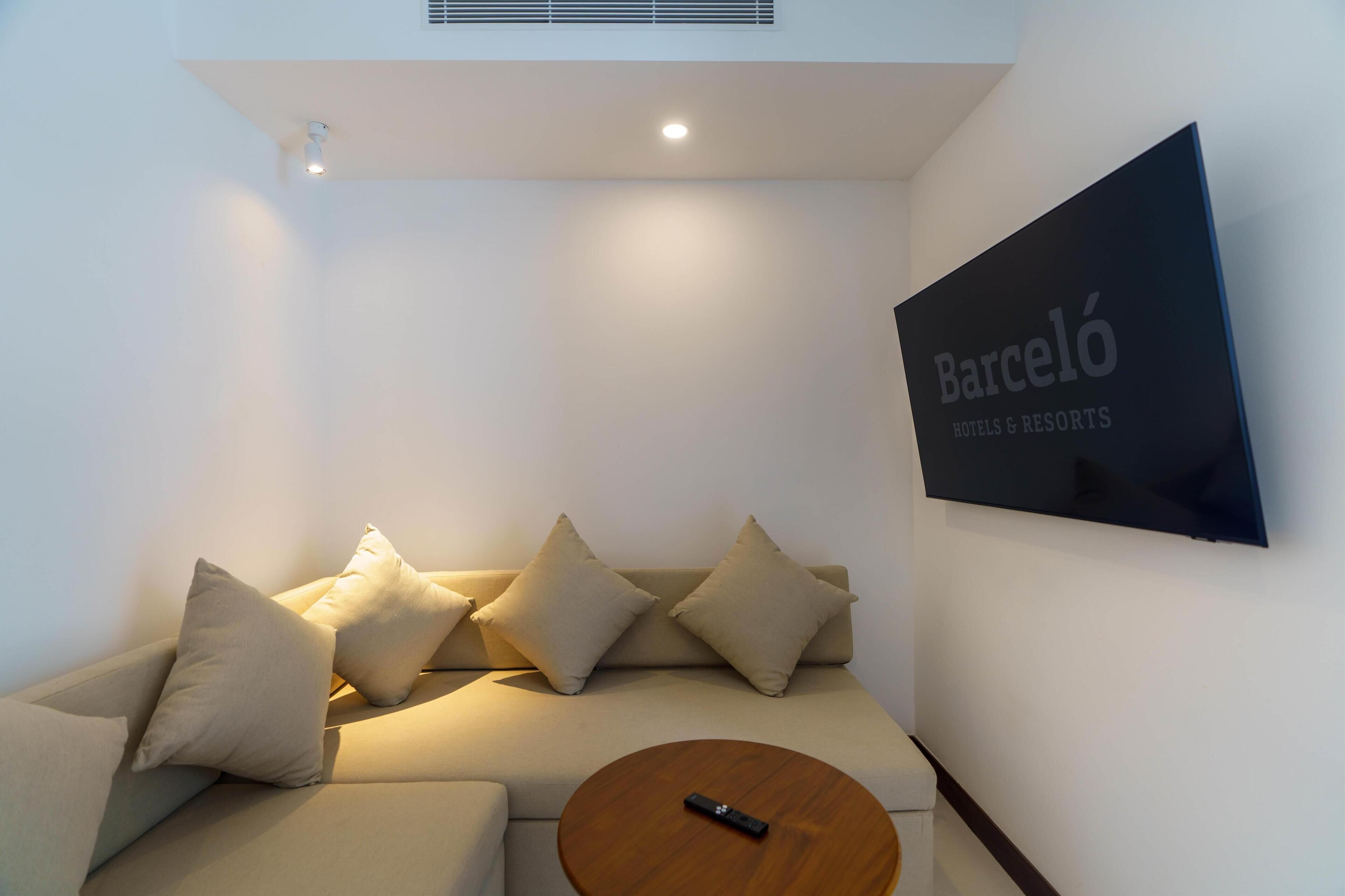 Een moderne woonkamer met een bank, kussens, een ronde salontafel en een tv met daarop 'Barceló Hotels & Resorts'.