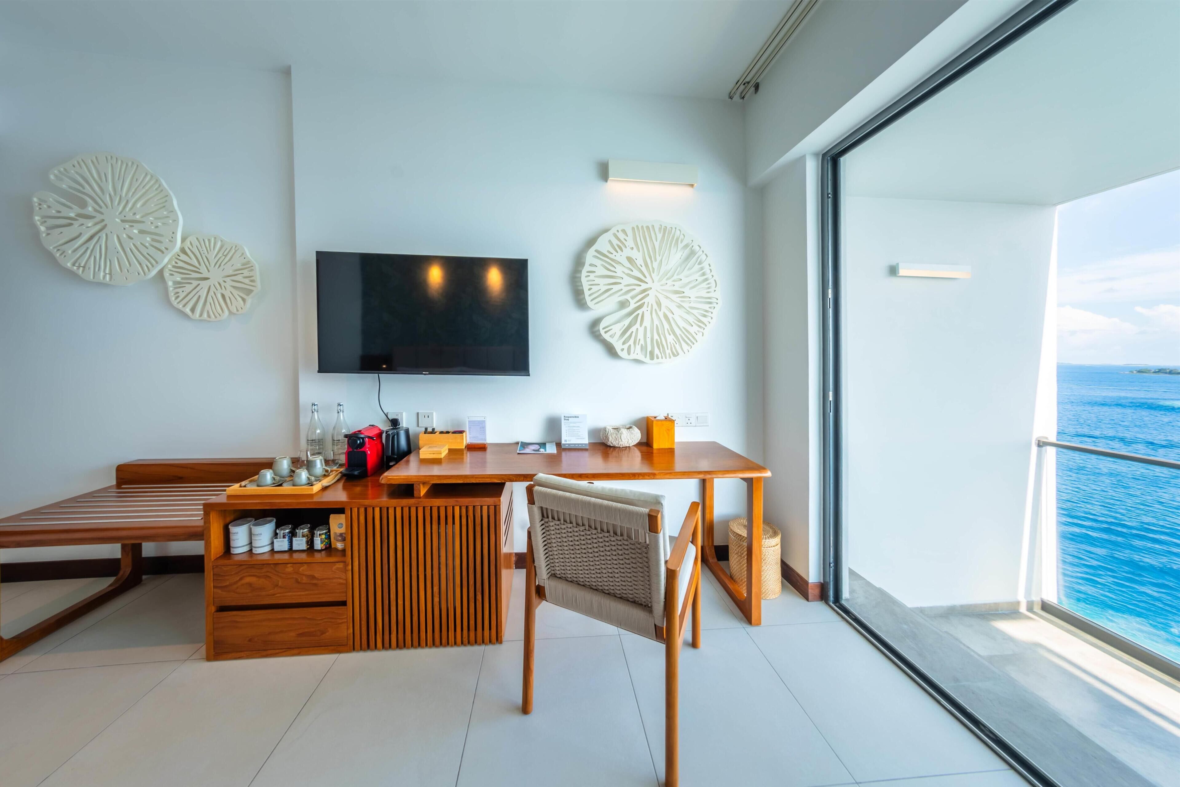 Une chambre moderne avec un bureau en bois, une télévision, des œuvres d’art décoratives au mur, un banc et une vue sur la mer.