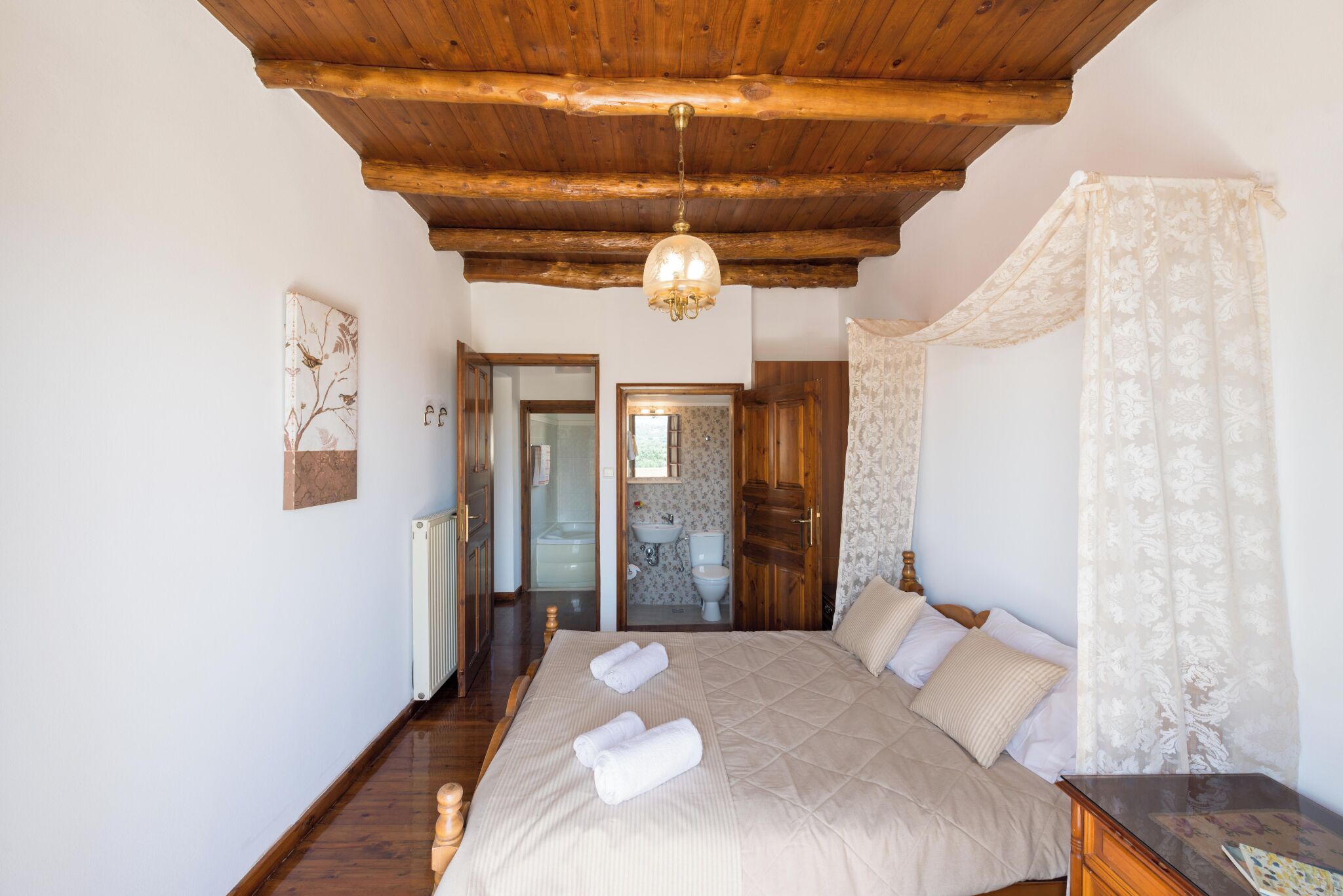Une chambre avec un plafond en bois, un grand lit recouvert d’une courtepointe, une table de chevet et une fenêtre ornée de rideaux légers.
