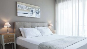 A neatly made bed with white linens and a grey headboard, flanked by two bedside tables with lamps, in a room with a large window and a wall-mounted artwork.