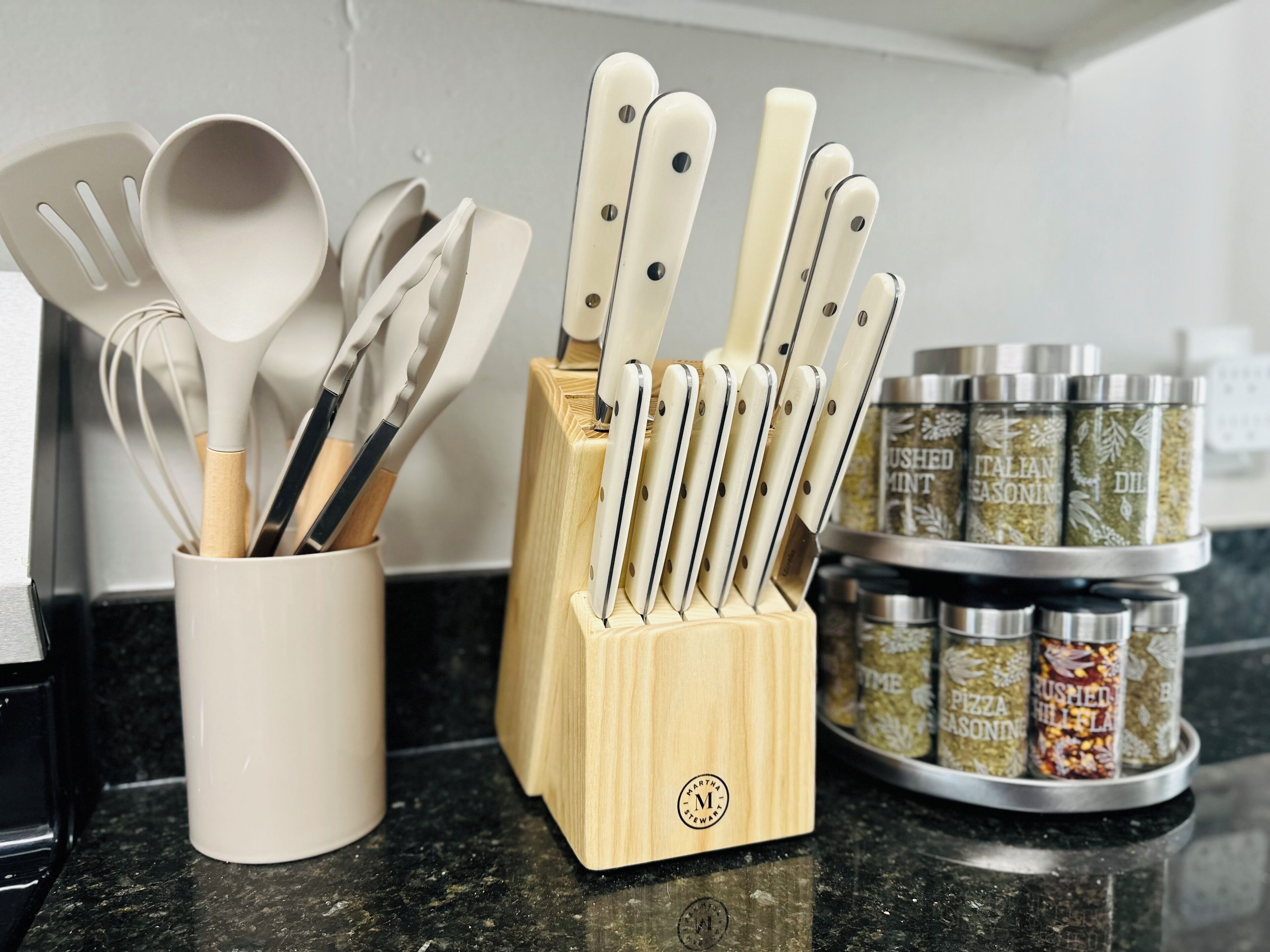 Una encimera de cocina con un portalapilas que contiene cuchillos y cucharas, una taza blanca y frascos de especias.