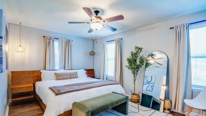 A modern bedroom with a wooden bed, a bench, a ceiling fan, a mirror, and a window with curtains. - Midtown Urban Retreat- Family Friendly 3BR Retreat (Tallahassee)