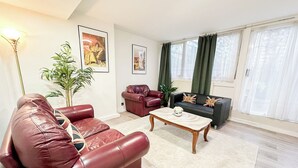 A living room with a red leather sofa, a black sofa with British flag cushions, a wooden coffee table, and a large window with sheer curtains.