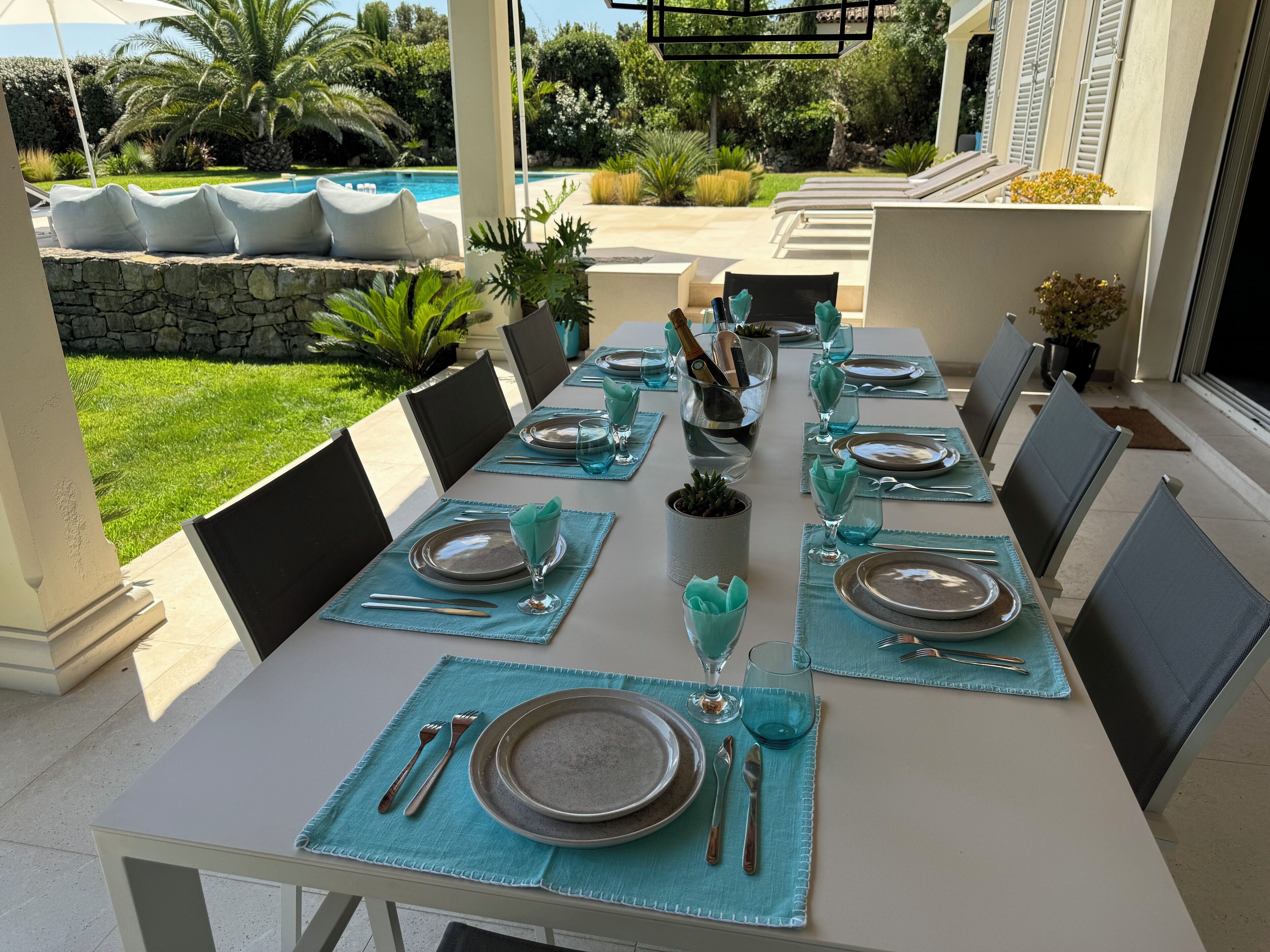 A dining area with a table set for six, surrounded by chairs, with a pool and greenery visible in the background.
