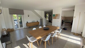 Un comedor moderno con una mesa de madera y sillas blancas, una cocina al fondo, y un ventanal que proporciona luz natural.