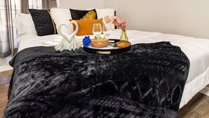 A neatly made bed with a black velvet bedspread, white pillows, and a tray with a heart-shaped towel, a vase with pink flowers, and a basket with a drink.