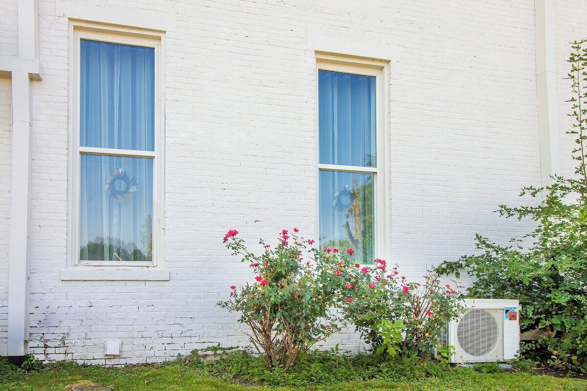 Un edificio bianco con due finestre, un piccolo arbusto con fiori rosa e un condizionatore d'aria.