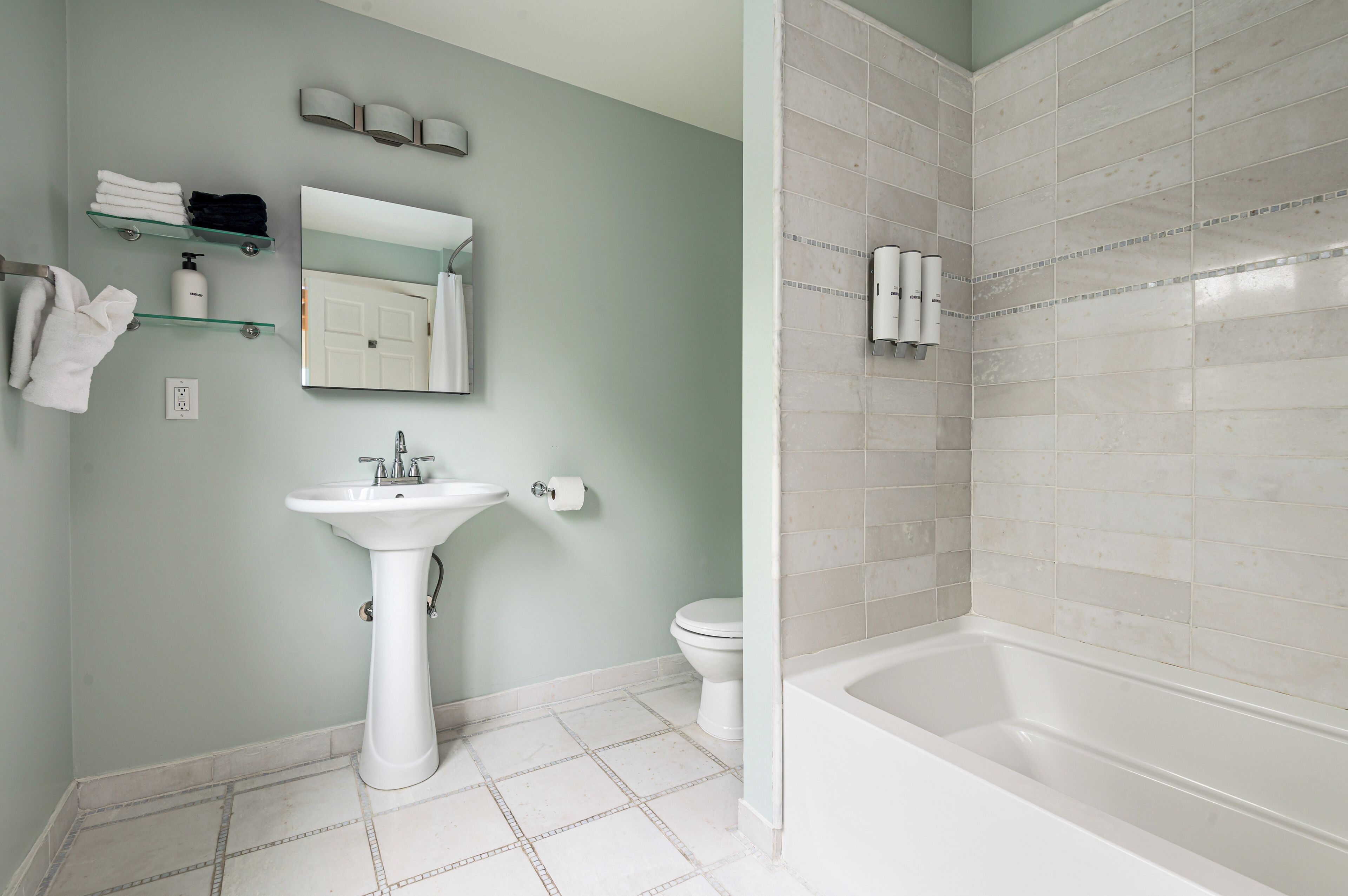 A bathroom with a pedestal sink, toilet, and bathtub. There are shelves with towels and toiletries, and a mirror above the sink.