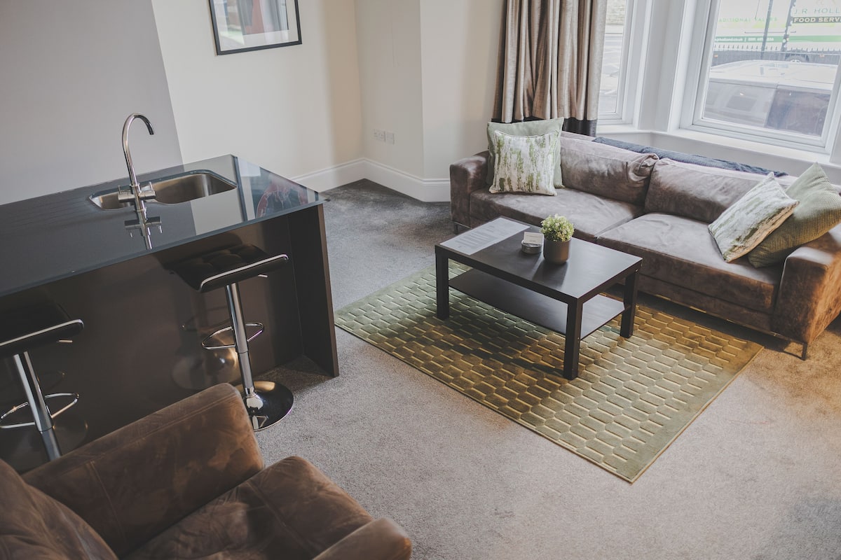 Ein modernes Wohnzimmer mit Küchenbereich, einem braunen Ledersofa, einem Couchtisch und Blick ins Freie durch große Fenster.