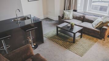 A modern living room with a kitchen area, a brown leather sofa, a coffee table, and a view of the outside through large windows.