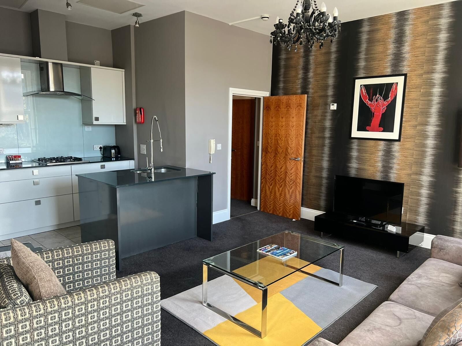 Una sala de estar moderna con una mesa de centro de vidrio, un sofá, un televisor y una zona de cocina con estufa y armarios.