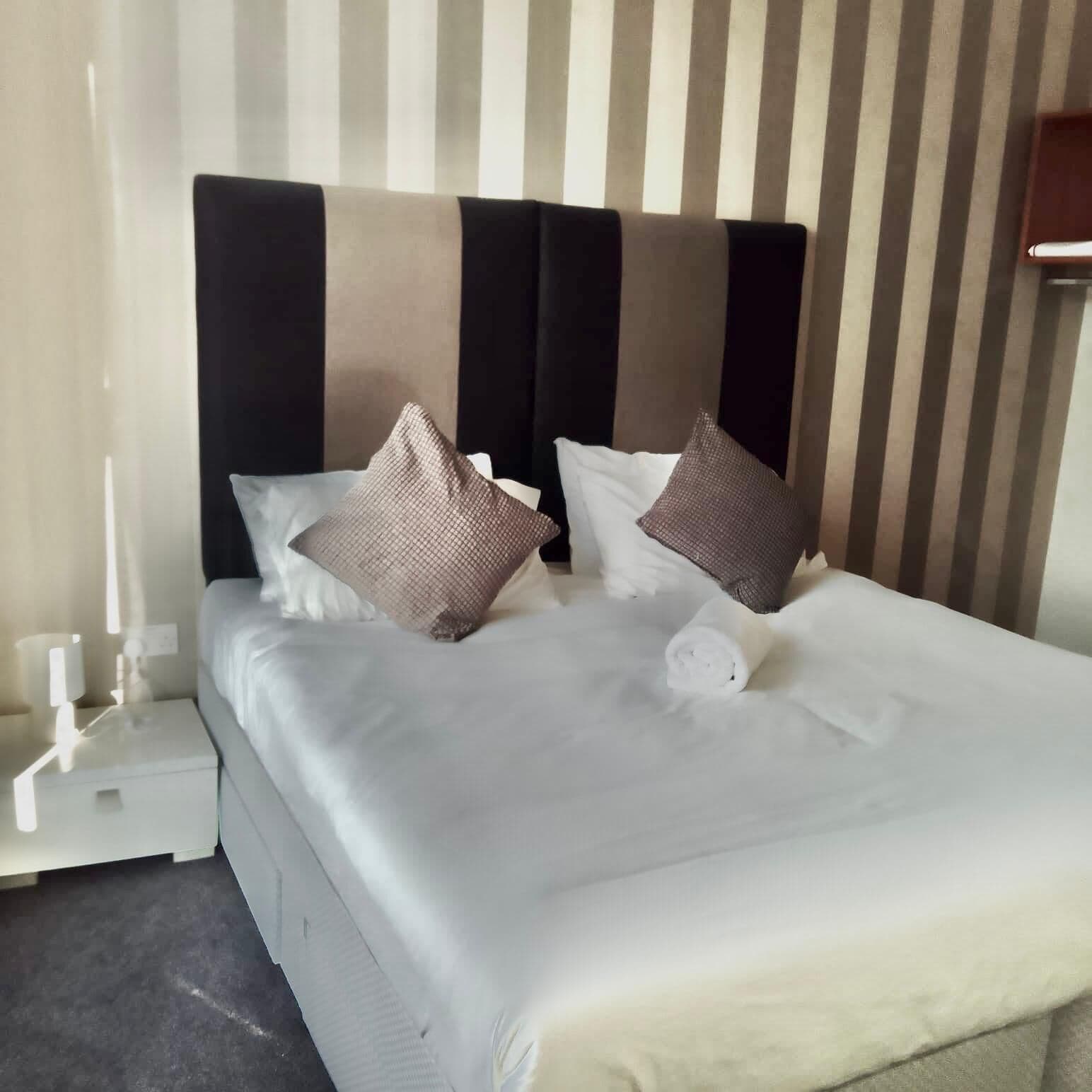 A neatly made bed with white linens and two brown pillows, a bedside table with a lamp, and a striped wall in the background.