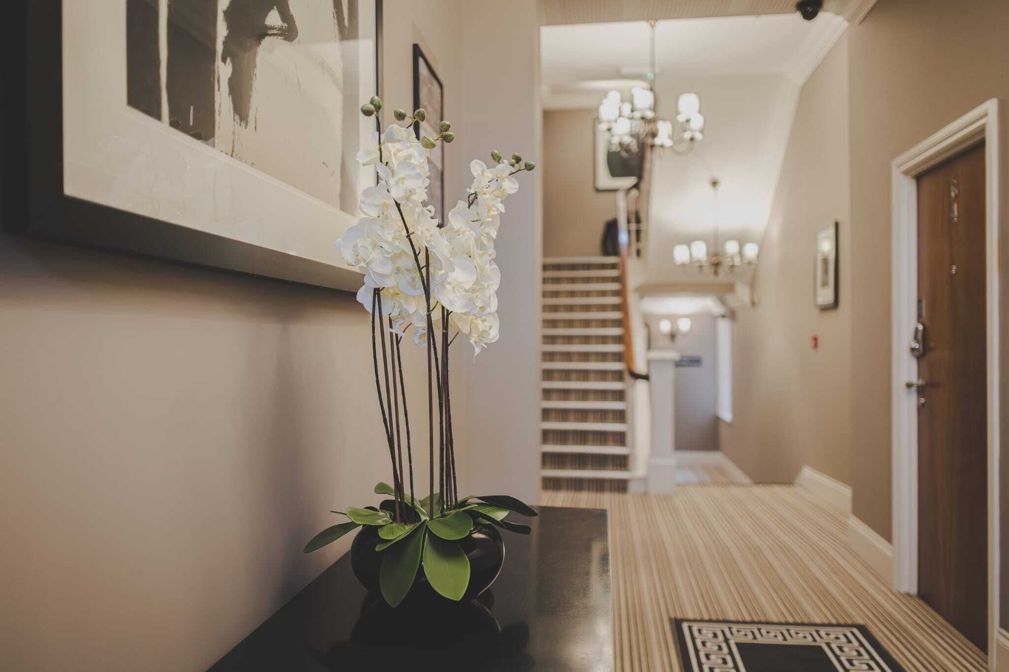 A hallway with a black vase of white orchids, a framed picture on the wall, and a staircase in the background.