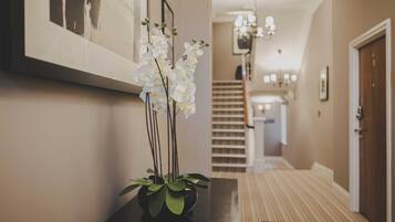 A hallway with a black vase of white orchids, a framed picture on the wall, and a staircase in the background.