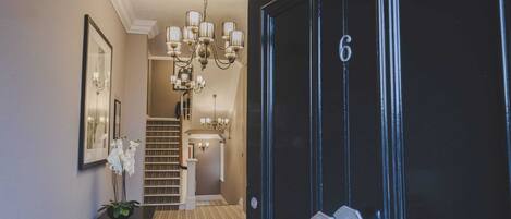 Un pasillo con una puerta negra numerada con el 6, una escalera y un candelabro.