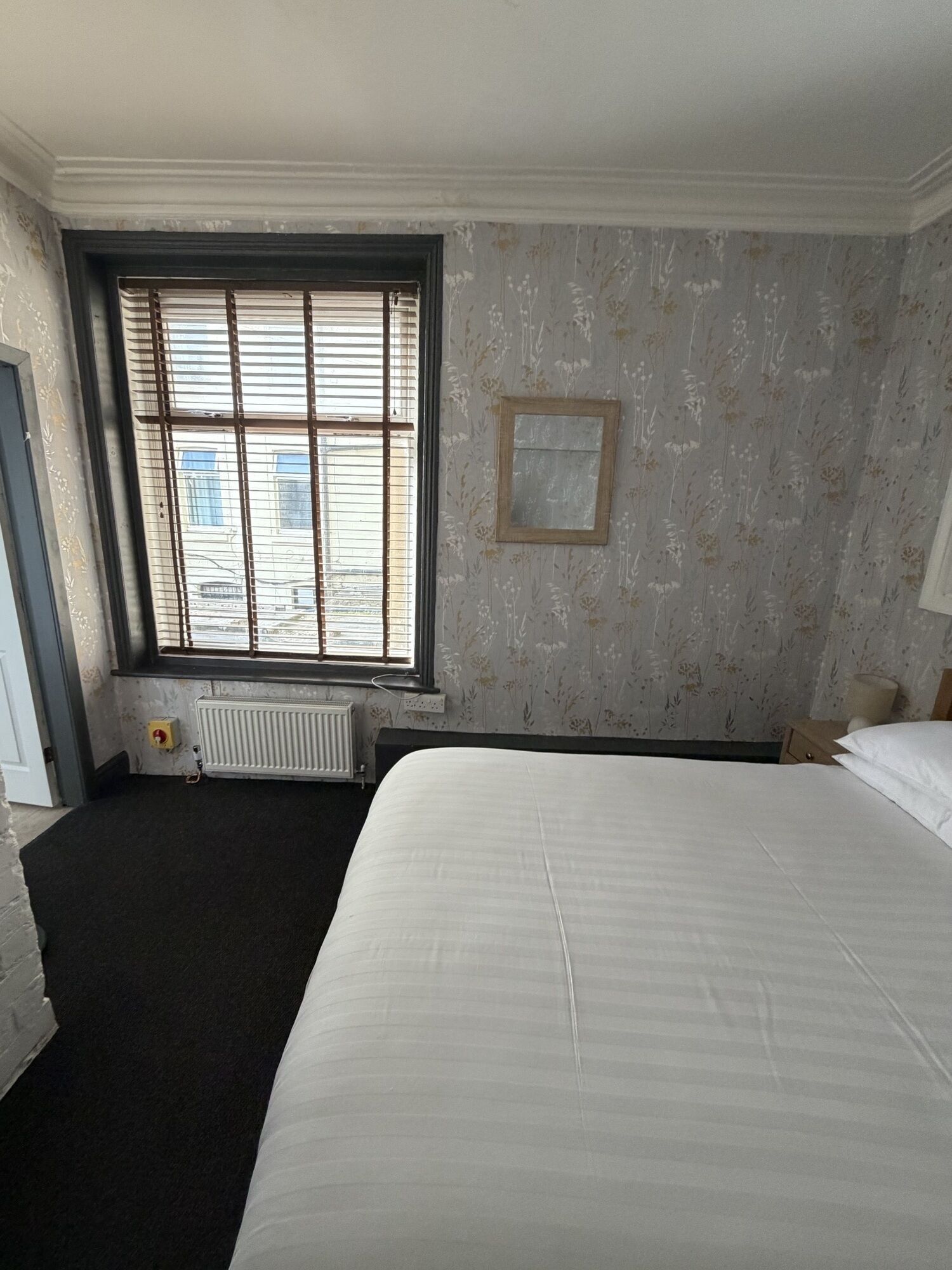 A hotel room with a large bed, a radiator, a window with blinds, and a wall-mounted picture.