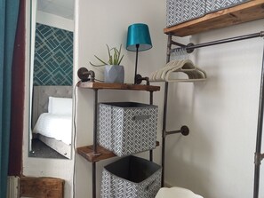 Ein kompakter Raum mit einem Bett, einem kleinen Regal mit Lampe, einem Blumentopf und einem Möbelstück mit Muster.